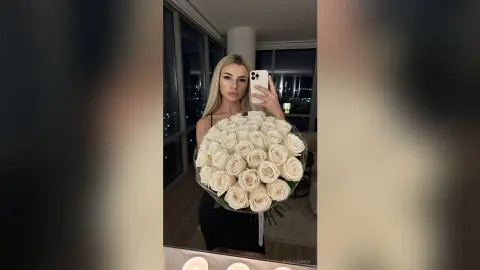 Blonde woman in a black dress holding a large bouquet of white roses in a modern apartment at night.