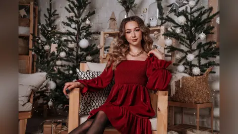Woman in a red velvet dress sitting in a cozy Christmas-themed room with decorated evergreen trees.