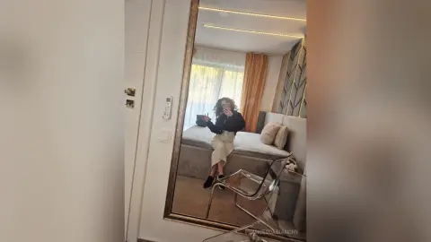 Young woman in a black top and beige skirt taking a mirror selfie in a modern, bright bedroom.