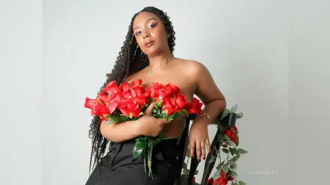 Beautiful woman with long braids holding a large bouquet of red roses in a minimalist studio setting.