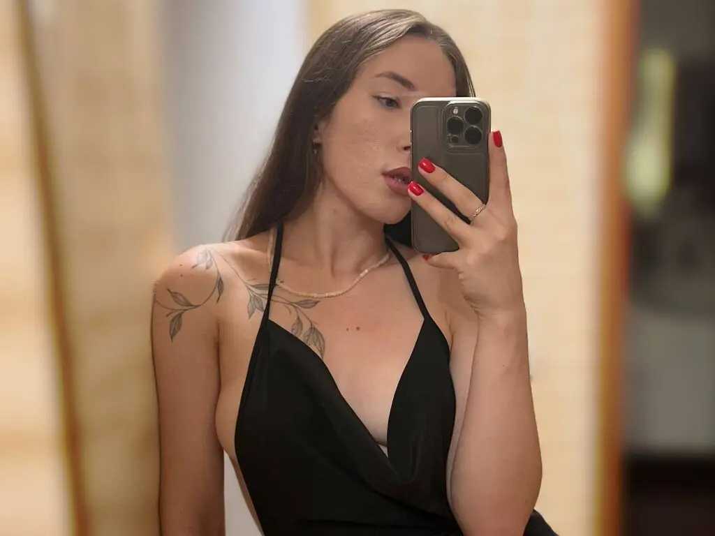 Young woman with long hair in a black halter dress taking a mirror selfie in a warm indoor setting.