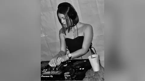 Young woman wearing headphones and a black top mixing music on a Pioneer DJ controller in a studio setting.