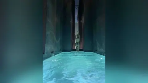Slim woman in a striped bikini relaxing in a luxury indoor turquoise pool within a moody, modern architectural space.