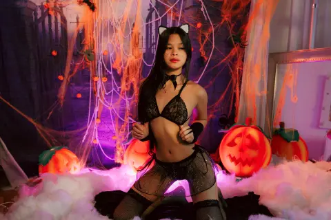 Woman in black lace cat costume sitting amidst glowing pumpkins and cobwebs in a spooky Halloween setting.