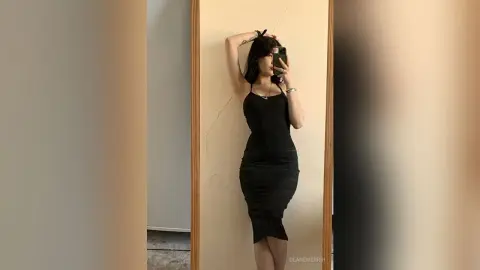 DeaneMerrih poses in a tight black tank dress, holding her phone up to a mirror in a room with light-colored walls.