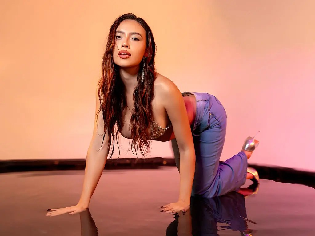 Glamorous woman in blue trousers posing on a reflective floor against a warm pink studio background.