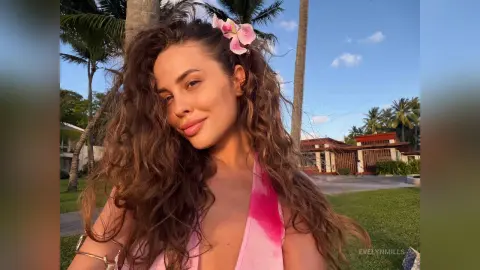 Smiling woman with wavy hair in a pink summer outfit enjoying a sunny tropical vacation at a palm tree resort.