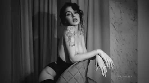 Moody black and white portrait of a tattooed woman sitting in a dimly lit room.