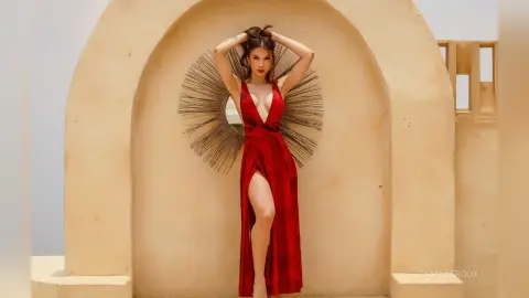 Elegant woman in a long red evening gown posing in a sunlit Mediterranean archway setting.