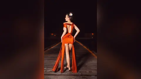 Elie poses on a wooden pier at night wearing a backless orange silk dress that features a high slit showing her bare legs.
