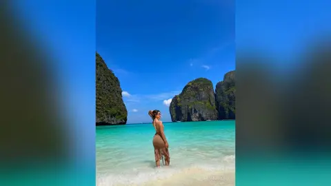 Woman in a tan swimsuit standing on a tropical beach with turquoise water and limestone cliffs under a blue sky.