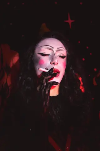 Woman with pale gothic makeup and dark wavy hair smoking a cigarette in a moody, red-lit cinematic setting.