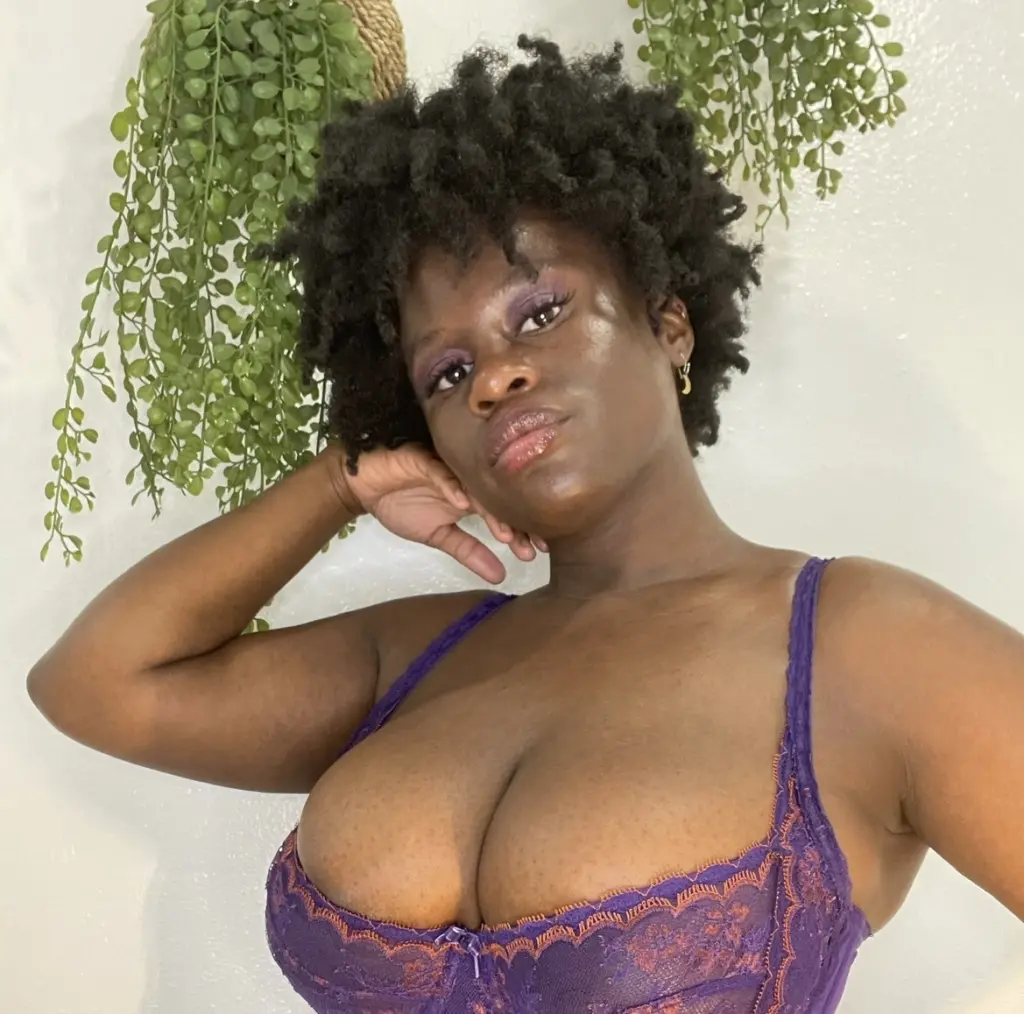 LaDiosa_Selene poses with her hands near her face, showing her dark curly hair and purple top against a leafy green background.