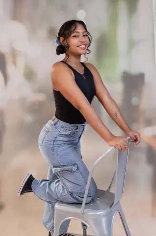 Smiling young woman in a black tank top and light blue denim jeans posing on a silver chair in a soft-focus setting.