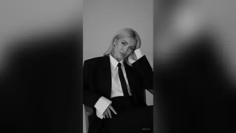 Anelise poses in a black suit and white shirt with a black tie, running her hand through her blonde hair against a plain background.