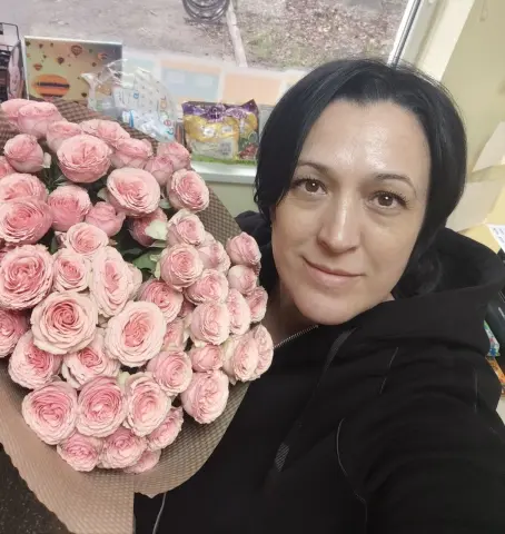 Smiling woman in a black hoodie holding a large bouquet of pink roses in a bright indoor setting.