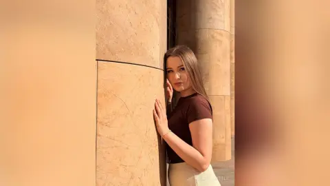 Young woman in a brown t-shirt leaning against a stone pillar in warm sunlight.