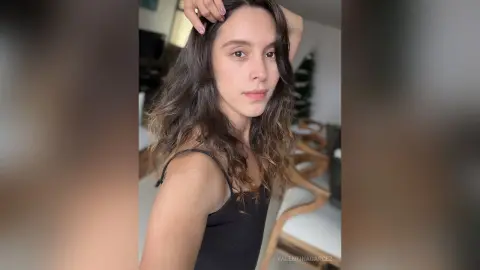 Young woman with long wavy hair wearing a black tank top in a bright, minimalist indoor setting.