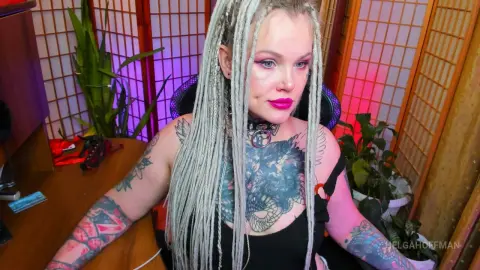 Tattooed woman with long blonde dreadlocks wearing a black top in a room with pink lighting and bamboo screens.