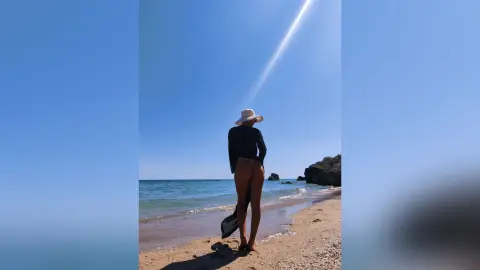 Woman in black swimsuit and white sun hat standing on a sunny beach looking at the blue ocean waves.
