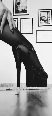 Elegant woman in black tights and high platform heels sitting near framed wall art in a moody black and white setting.