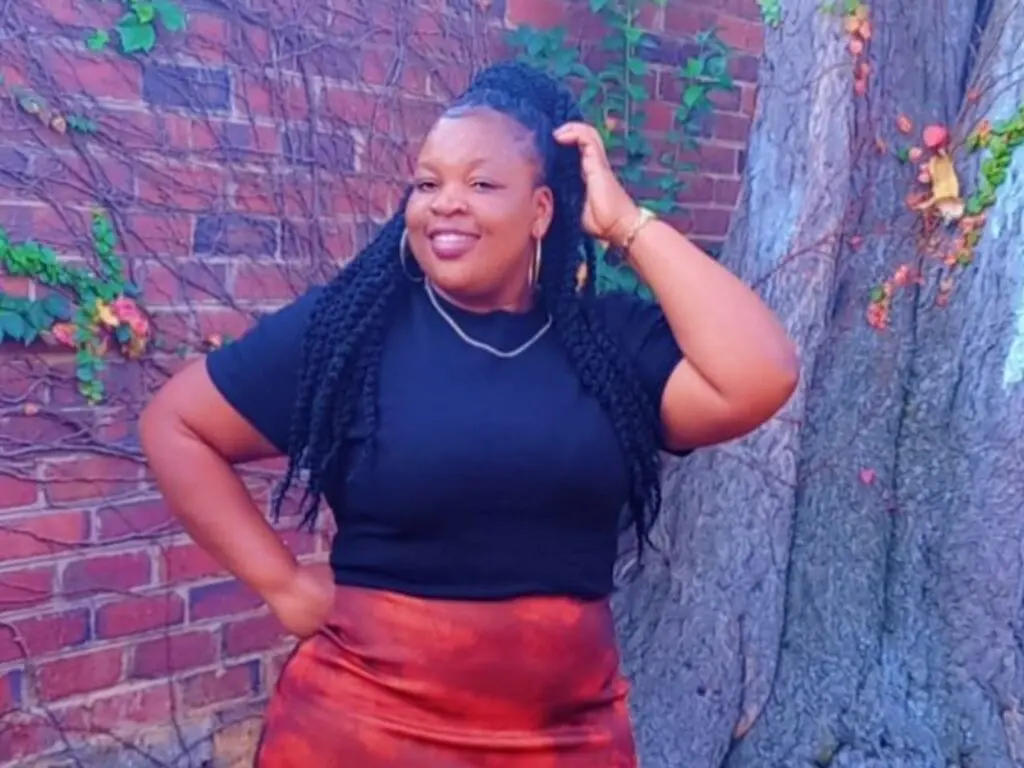 PearlyBrown smiles while posing outdoors in a black t-shirt and red skirt, with her long black braids pulled back and one hand touching her hair.