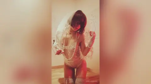 Romantic woman in white lace lingerie and veil wearing pearls in a soft lit indoor setting.