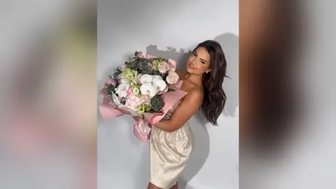 Smiling woman in a champagne satin dress holding a large bouquet of pink and white flowers against a plain wall.