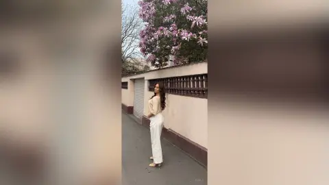 Stylish woman in a cream blouse and white trousers standing by a flowering pink tree on a city street.