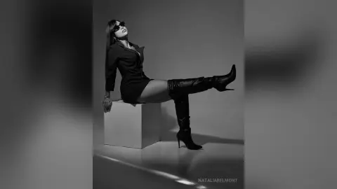 Stylish woman in a black dress and tall boots posing confidently on a white pedestal in a studio setting.