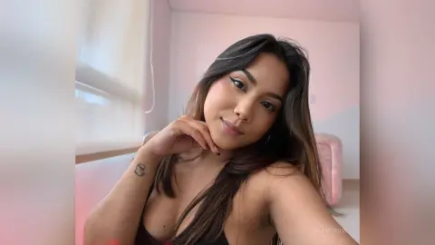 Young woman with long brown hair and winged eyeliner posing in a bright, cozy room.