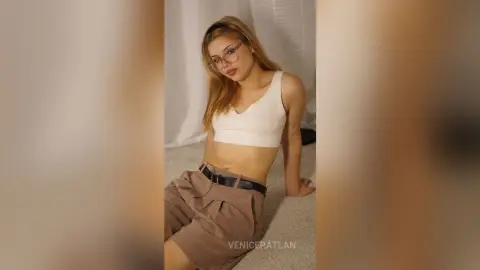 Young woman in white crop top and tan trousers posing in a bright, minimalist indoor setting.