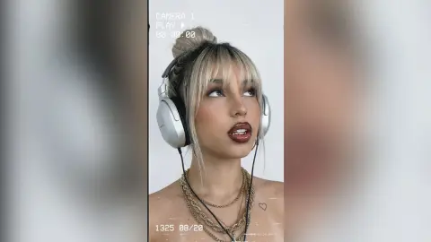 Blonde woman with bangs wearing white headphones and gold necklaces in a soft, cinematic indoor setting.
