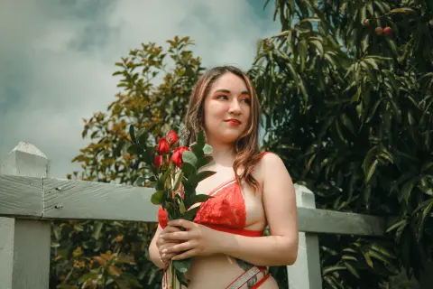 Young woman in red lace lingerie holding a bouquet of red roses in a lush green garden setting.