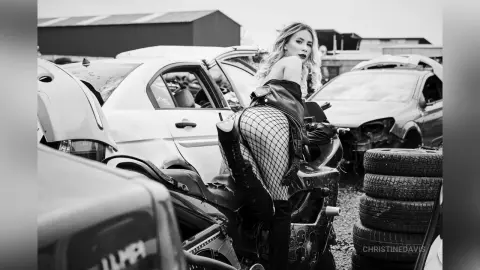 ChristineDavis poses in a junkyard wearing a black leather jacket and fishnet stockings, leaning against a white car.