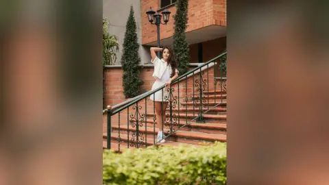 TefyRusso poses on outdoor brick stairs wearing a white sleeveless top and white skirt, leaning against a black metal railing with her hand behind her head.