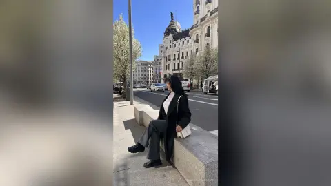 NashiroSakurai sits on a stone ledge on a city street wearing a long black coat and grey trousers.