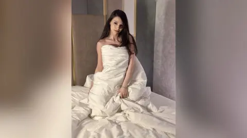 Elegant woman in white silk sheets relaxing on a bed in a bright, dreamy bedroom setting.