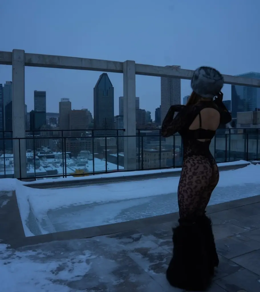 Elliepixiee poses outdoors in a long-sleeve black lace bodysuit, showing her long brown hair and figure against a snowy balcony setting.
