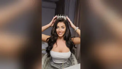 Smiling woman in a white tank top wearing a silver crystal tiara in a bright indoor setting.