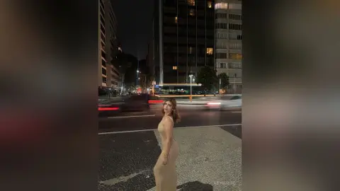 Elegant woman in a beige cocktail dress standing on a city street at night near glowing urban buildings.