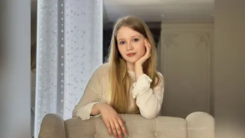 Young woman in a white long-sleeve blouse relaxing on a beige sofa in a bright, airy room.