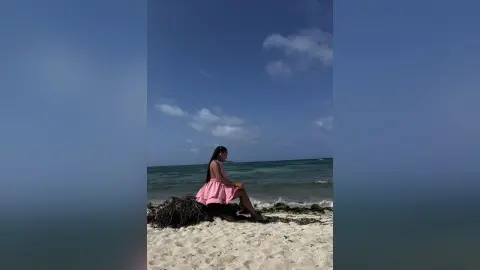 Young woman in a pink summer dress sitting on a sandy beach by the ocean under a blue sky.