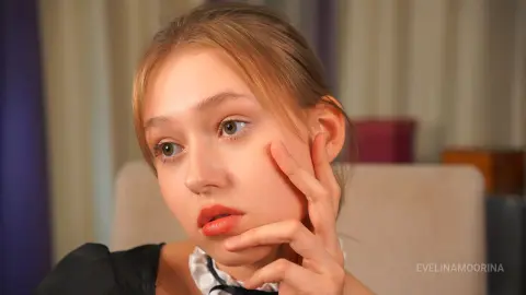 Pensive young woman with blonde hair in a black outfit sitting in a cozy indoor setting.