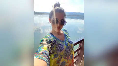 Woman in colorful patterned tunic and sunglasses smiling on a boat overlooking a calm lake at sunset.