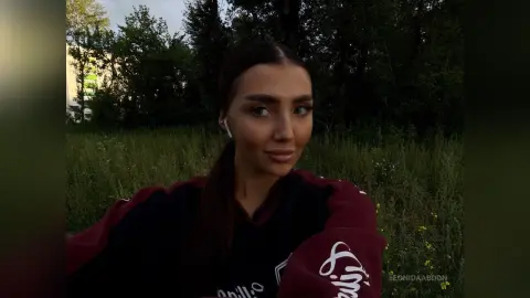 LeonidaAbdon poses outdoors in a field, wearing a dark maroon and black long-sleeved top with her dark hair pulled back.