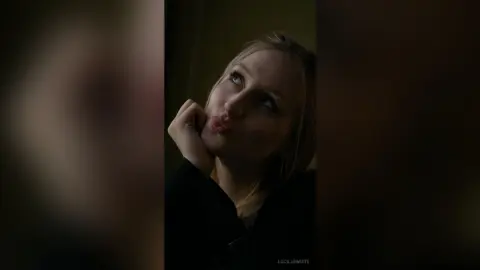 Dreamy portrait of a blonde woman in a black outfit looking upward in moody, low-light indoor setting.
