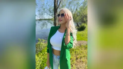 Blonde woman in a green blazer and white crop top smiling by a serene lake on a sunny day.