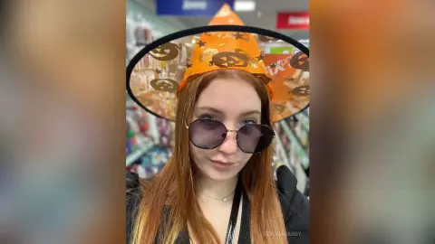 Redheaded woman in a festive orange witch hat and sunglasses shopping for Halloween decor in a store.