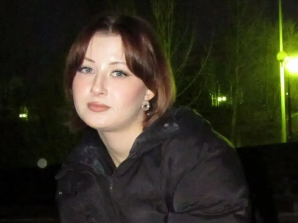 Young woman in a black winter jacket sitting outdoors at night under soft street lighting.